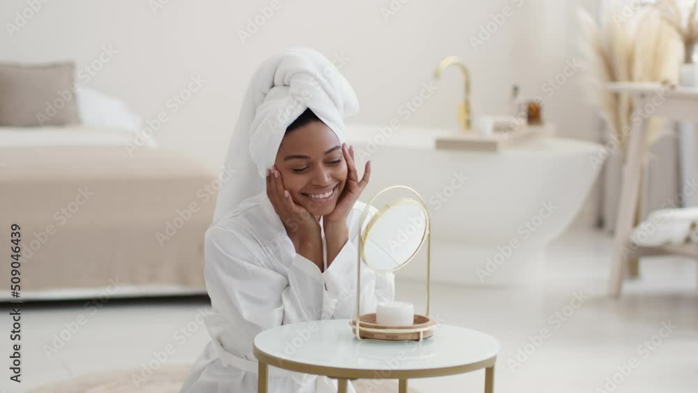 Young pretty african american woman with towel on head looking at mirror and touching face, massaging skin, slow motion Young pretty african american woman with towel on head looking at mirror and touching face, massaging skin, slow motion
