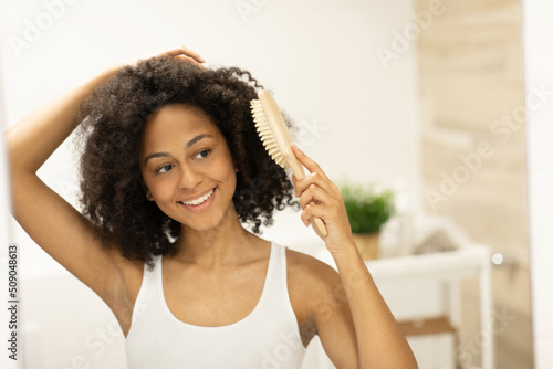 A young woman in a tank top stands in front of a bathroom mirror and combs her hair with a wooden brush after a shower.