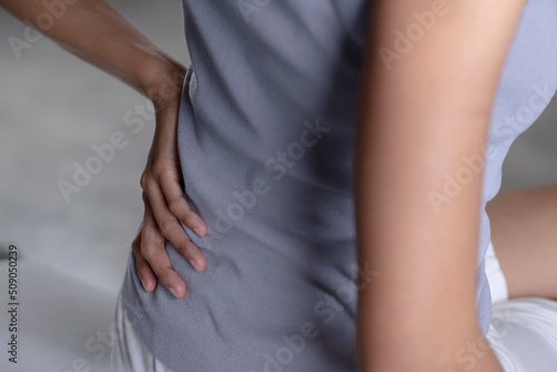 Young Asian woman sitting on bed and suffering from back pain. Adult female is holding her lower back and suffering from chronic back pain. Health care and medical concept.