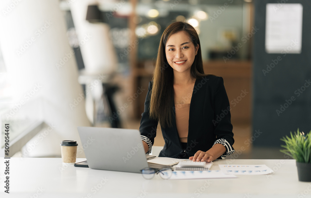 Fototapeta premium Cheerful business lady working on laptop in office, Asian happy beautiful businesswoman in formal suit work in workplace. Attractive female employee office worker smile.