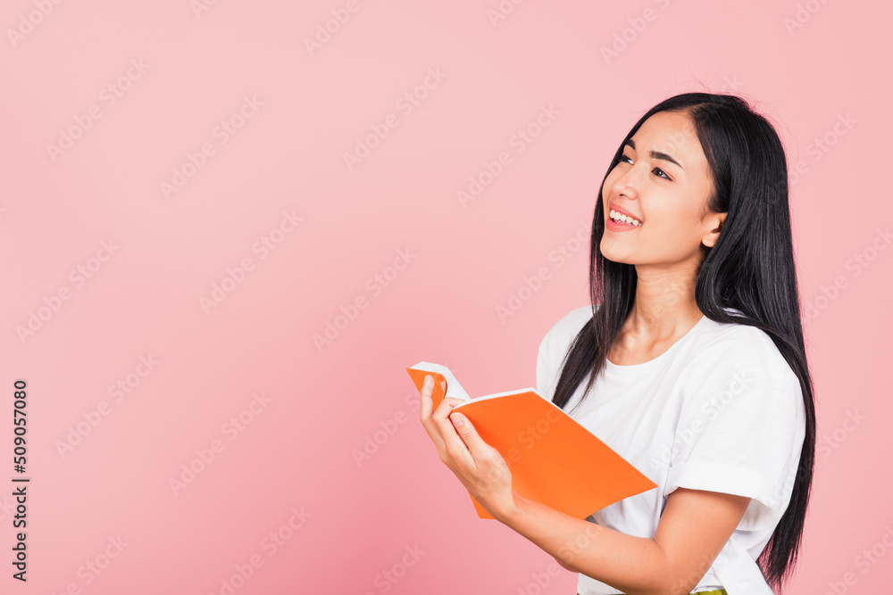 Portrait of happy Asian beautiful young woman confident smiling standing holding orange book open or diary for reading, studio shot isolated on pink background, with copy space, education concept Portrait of happy Asian beautiful young woman confident smiling standing holding orange book open or diary for reading, studio shot isolated on pink background, with copy space, education concept