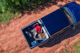 Aerial view of a woman sitting in the back of a pickup truck with her dog and suitcase, all on a typical red sand road in Paraguay.