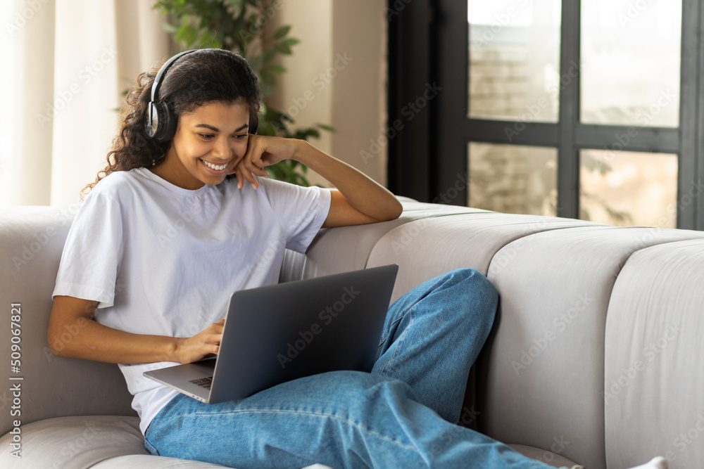 Young African American hipster woman in earphone with Afro hair using a laptop talking to webcam with friend online lying on the couch at home. Video call, virtual chat concept Young African American hipster woman in earphone with Afro hair using a laptop talking to webcam with friend online lying on the couch at home. Video call, virtual chat concept