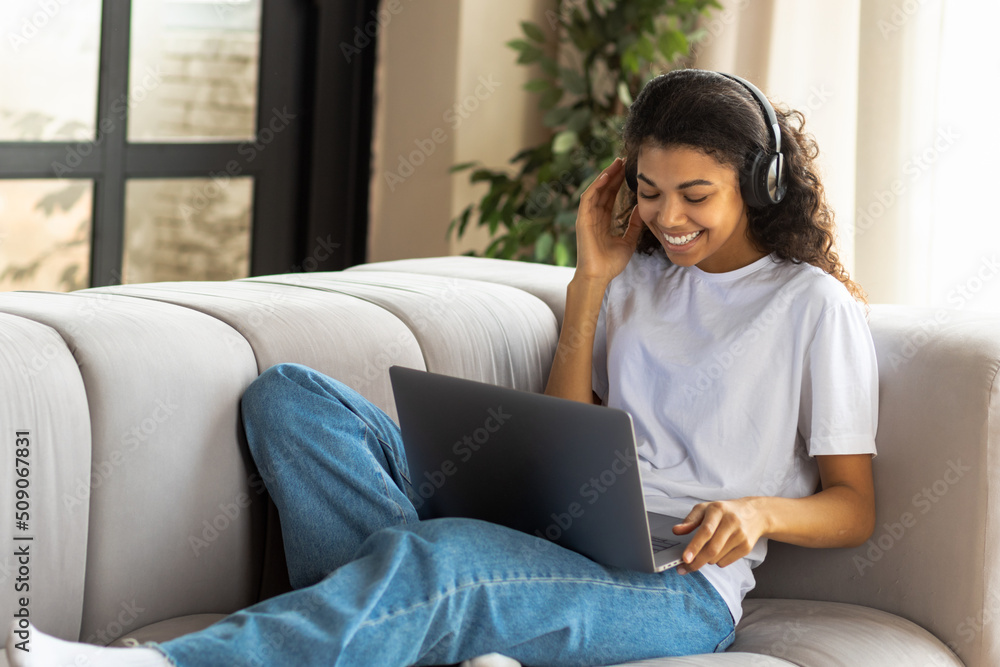 Video call, virtual chat concept. Young African American hipster woman in earphone with Afro hair using a laptop talking to webcam with friend online lying on the couch at home Video call, virtual chat concept. Young African American hipster woman in earphone with Afro hair using a laptop talking to webcam with friend online lying on the couch at home