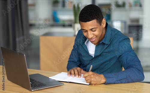 Portrait of confident African American young man. Successful student or office worker sitting at workplace and working using laptop, smiling friendly