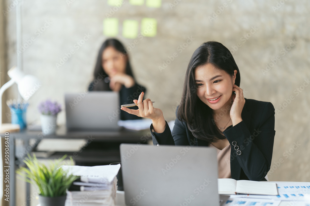 Attractive asian young confident businesswoman sitting at the office table with group of colleagues in the background, working on laptop computer. Attractive asian young confident businesswoman sitting at the office table with group of colleagues in the background, working on laptop computer.