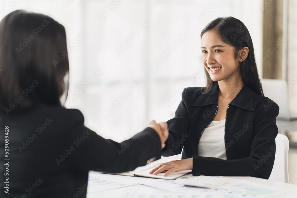 Two Businesswomen Shaking Hands In Modern Office, Smiling young businesswoman shaking hands with a coworker during a meeting with colleagues around a table in an office boardroom. Two Businesswomen Shaking Hands In Modern Office, Smiling young businesswoman shaking hands with a coworker during a meeting with colleagues around a table in an office boardroom.