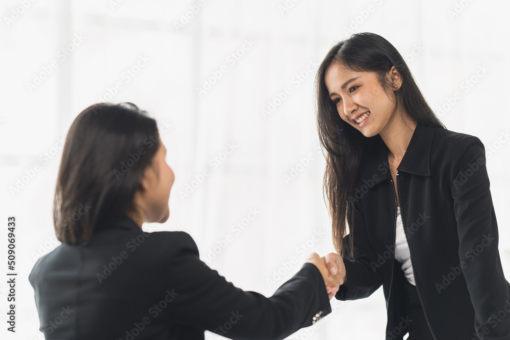 Two Businesswomen Shaking Hands In Modern Office, Smiling young businesswoman shaking hands with a coworker during a meeting with colleagues around a table in an office boardroom. Two Businesswomen Shaking Hands In Modern Office, Smiling young businesswoman shaking hands with a coworker during a meeting with colleagues around a table in an office boardroom.