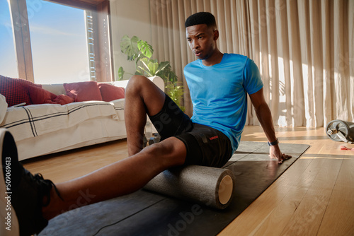 Young African American Black male stretching his hamstring using a foam roller on an exercise mat, working out at home in his living room