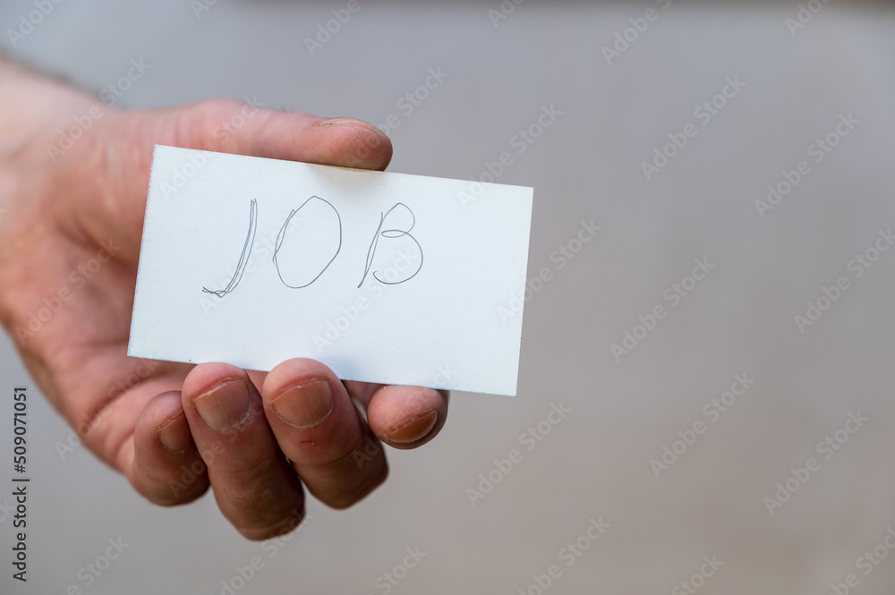 Job. A word written in jagged letters. A man's hand holds a white paper rectangle with text against a gray background. Job. A word written in jagged letters. A man's hand holds a white paper rectangle with text against a gray background.