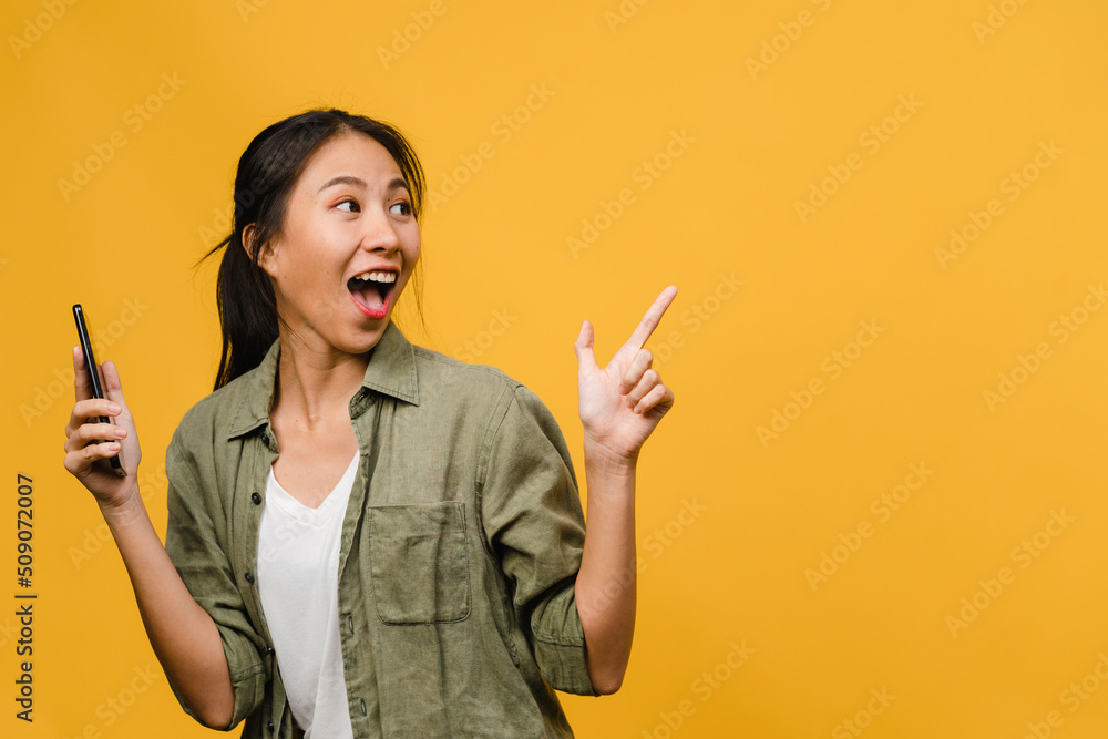 Portrait of young Asian lady using mobile phone with cheerful expression, show something amazing at blank space in casual clothing and stand isolated over yellow background. Facial expression concept. Portrait of young Asian lady using mobile phone with cheerful expression, show something amazing at blank space in casual clothing and stand isolated over yellow background. Facial expression concept.