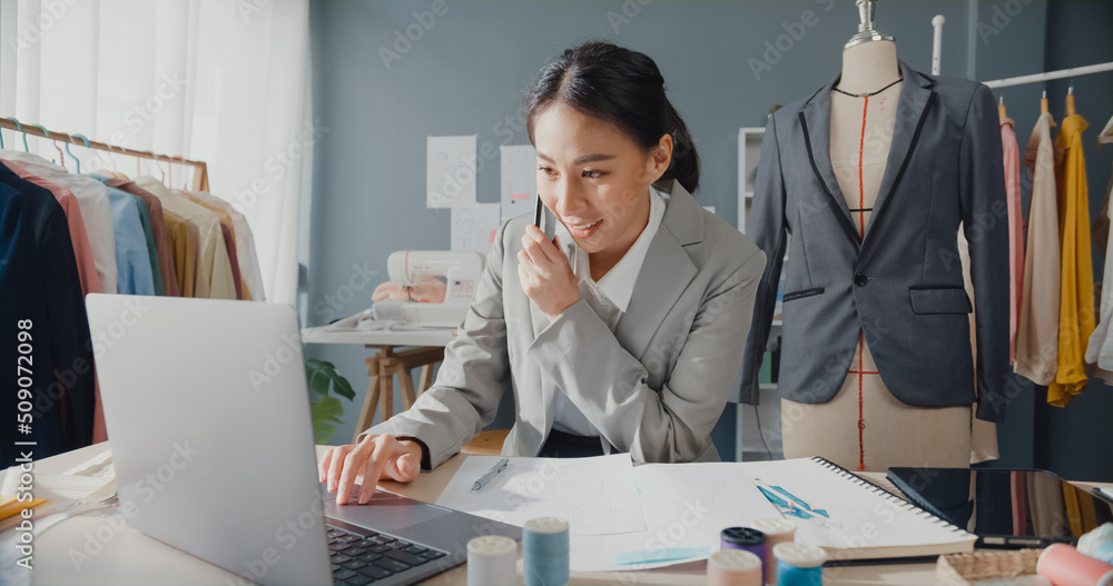 Professional cheerful Asia businesswoman designer sitting on desk use laptop computer call smartphone to customer confirm purchase order online marketplace at studio workplace. Successful business. Professional cheerful Asia businesswoman designer sitting on desk use laptop computer call smartphone to customer confirm purchase order online marketplace at studio workplace. Successful business.