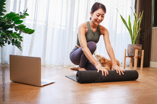 young Asian woman unrolling roll black yoga mat with her dog at home, New normal lifestyle concept