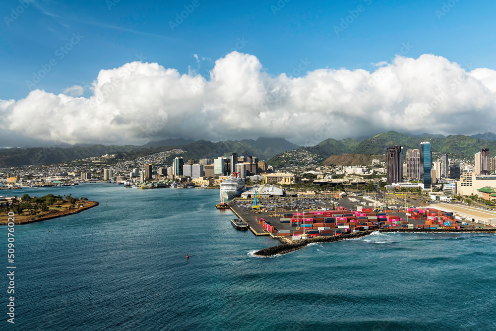 Naklejka premium Honolulu city and industrial zone with containers on the lot. Aerial view of industrial zone and tall buildings by the ocean. Clouds over the mountains in background