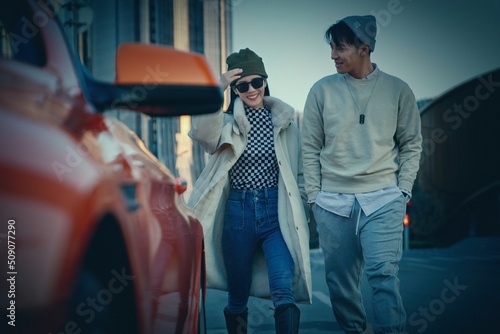 a Fashionable young couple with sunglasses and walking beside an orange luxury sports car photo