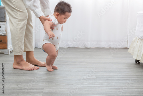 Asian baby taking first steps walk forward. Happy little baby learning to walk with mother help at home. Mother teaching how to walk gently. parenthood, family,baby growth and development concept