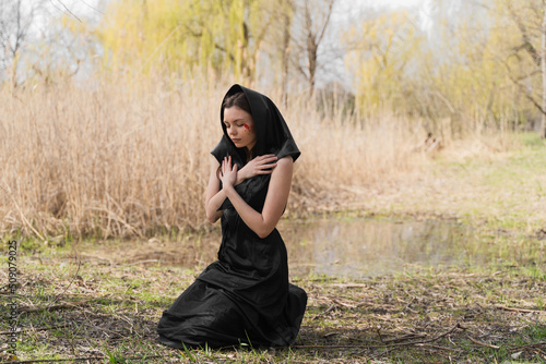 A young widow in a black mourning dress and headscarf kneeling on the ground, praying and crying with bloody tears.