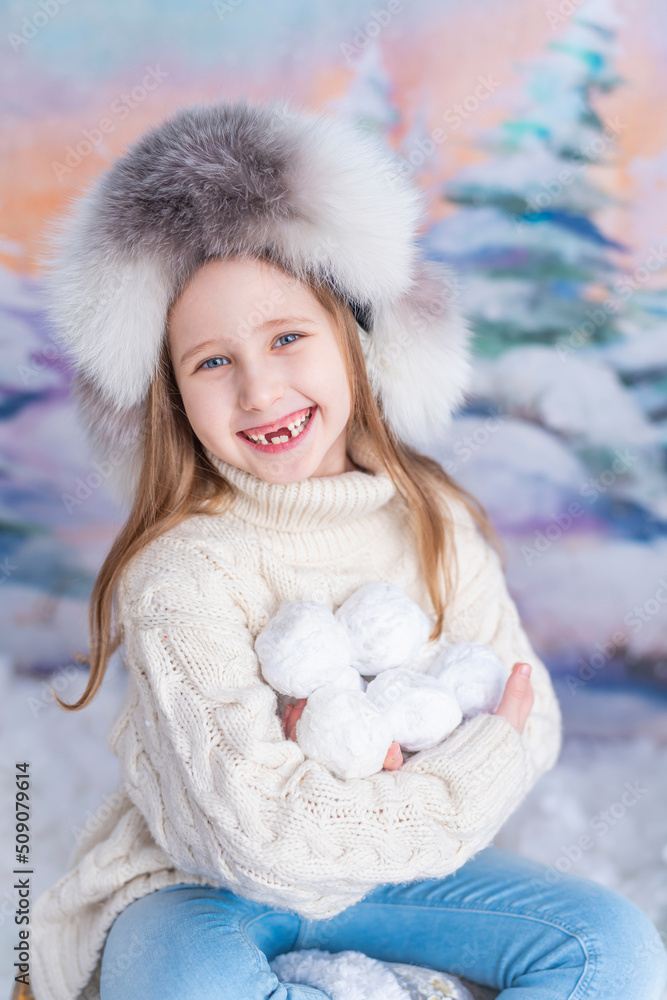 happy blue-eyed baby girl in a fur hat and a knitted sweater, holding a pile of snowballs, snowballs, sitting against the background of a winter forest. Winter holidays, warm clothes, winter game happy blue-eyed baby girl in a fur hat and a knitted sweater, holding a pile of snowballs, snowballs, sitting against the background of a winter forest. Winter holidays, warm clothes, winter game