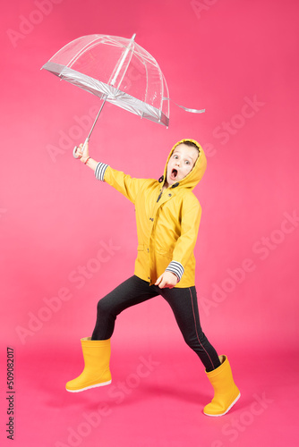 Happy little girl wearing yellow raincoat and playing with umbrella isolated on pink background. Smiling to bad weather concept.