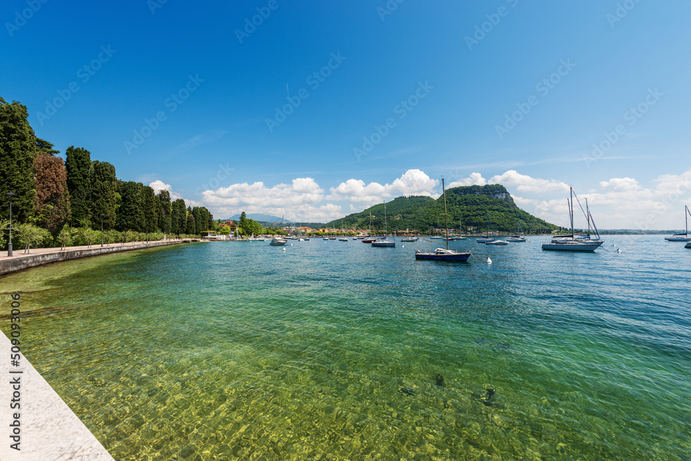 Obraz premium Beautiful bay of Lake Garda with many boats moored in front of the small town of Garda, tourist resort on the coast of Lake Garda. On the horizon the hill, Rocca di Garda. Verona, Veneto, Italy.
