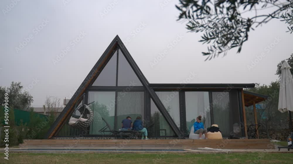 People sit on bench and chair near modern triangle-shaped country house with panoramic windows. Family relaxes in yard on cloudy day People sit on bench and chair near modern triangle-shaped country house with panoramic windows. Family relaxes in yard on cloudy day