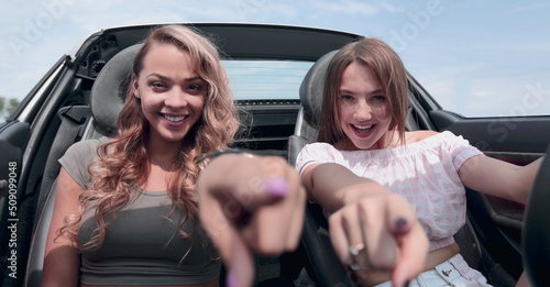 two happy young women sitting in a car and pointing at you. © ASDF