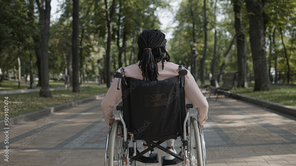 Back view of African American woman with disability riding wheelchair along park, resting outdoor