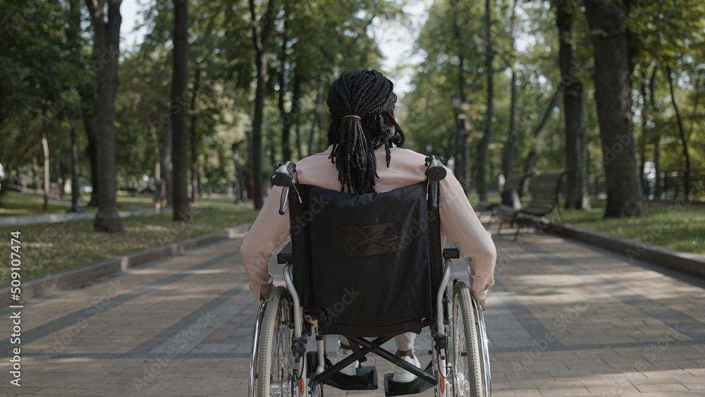 Obraz premium Back view of African American woman with disability riding wheelchair along park, resting outdoor