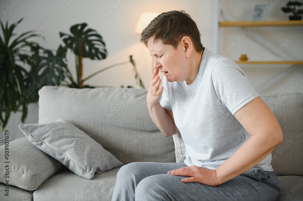 Sick adult woman coughing covering mouth with tissue sitting on a couch at home.