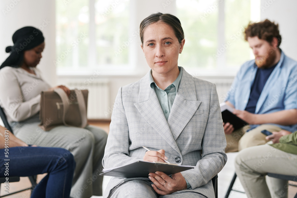 Young confident female counselor in elegant grey suit holding document while sitting in front of her patients and looking at camera Young confident female counselor in elegant grey suit holding document while sitting in front of her patients and looking at camera