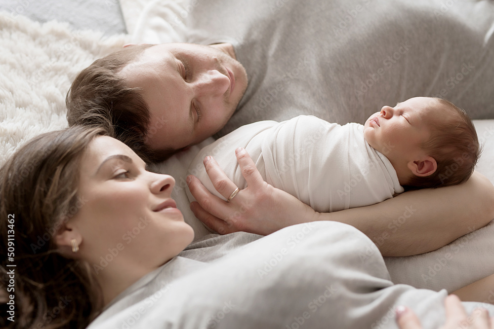 Newborn baby in mother's and father's arms on bed. Postcard Mother's and father's day. Children Protection Day. World Happiness Day. Smiling child. Happy parenthood Newborn baby in mother's and father's arms on bed. Postcard Mother's and father's day. Children Protection Day. World Happiness Day. Smiling child. Happy parenthood