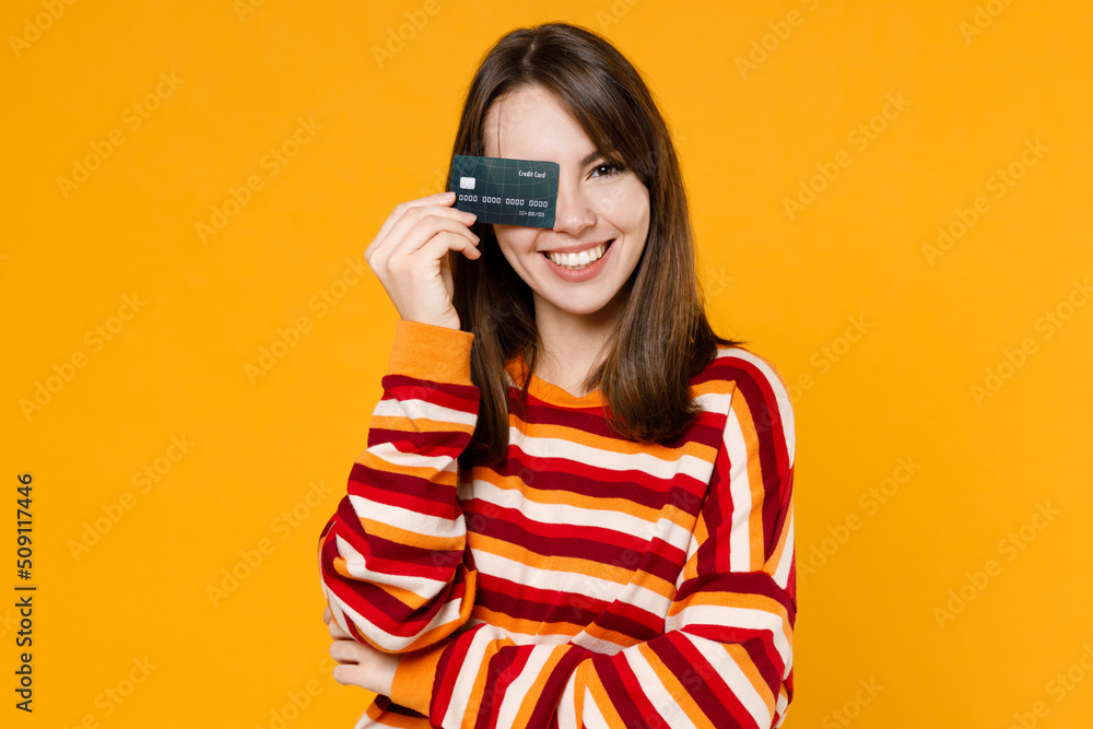 Young happy smiling cheerful excited satisfied woman 20s in red striped sweatshirt hold cover eye with credit bank card isolated on plain yellow background studio portrait. People lifestyle concept. Young happy smiling cheerful excited satisfied woman 20s in red striped sweatshirt hold cover eye with credit bank card isolated on plain yellow background studio portrait. People lifestyle concept.