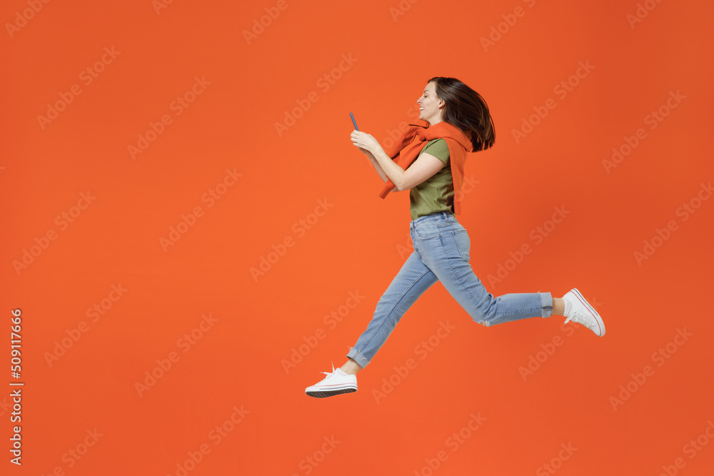 Full body side view young smiling happy woman 20s in khaki t-shirt tied sweater on shoulders jump high hold in hand use mobile cell phone isolated on plain orange background. People lifestyle concept.