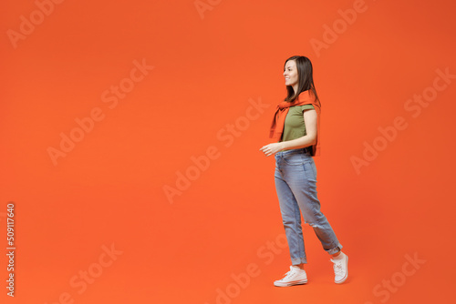Full body side view young smiling happy woman 20s wearing khaki t-shirt tied sweater on shoulders looking asidewalk go stroolling isolated on plain orange background studio. People lifestyle concept.