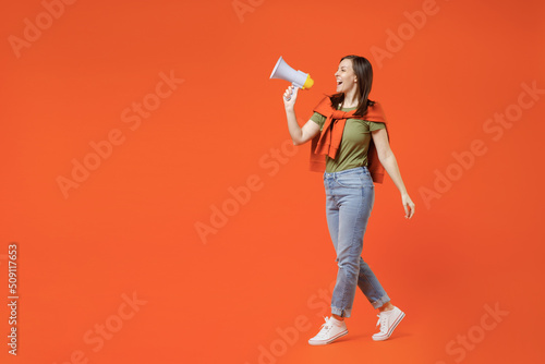 Full body side view young happy woman 20s wear khaki t-shirt tied sweater on shoulders hold scream in megaphone announces discounts sale Hurry up isolated on plain orange background studio portrait