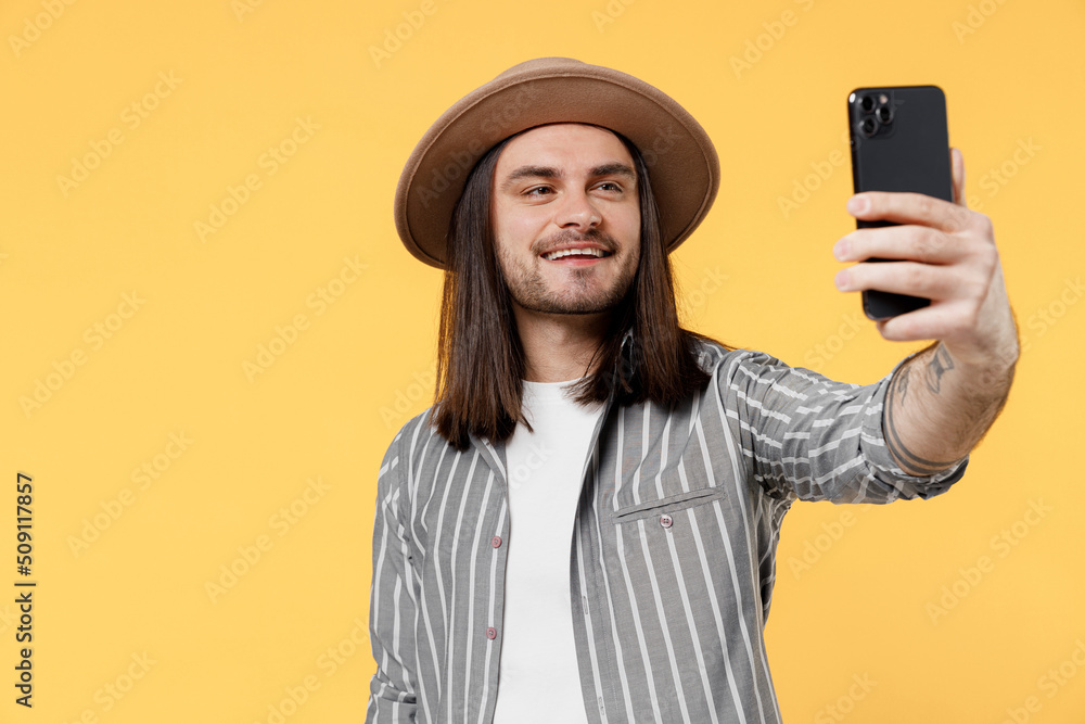Young fun man he 20s wears striped grey shirt white t-shirt hat doing selfie shot on mobile cell phone post photo on social network isolated on plain yellow color background. People lifestyle concept. Young fun man he 20s wears striped grey shirt white t-shirt hat doing selfie shot on mobile cell phone post photo on social network isolated on plain yellow color background. People lifestyle concept.