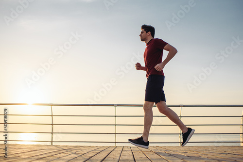 Side view full size young strong sporty athletic fit sportsman man in sports clothes jogging jump high warm up training at sunrise sun dawn over sea beach outdoor on pier seaside in summer day morning