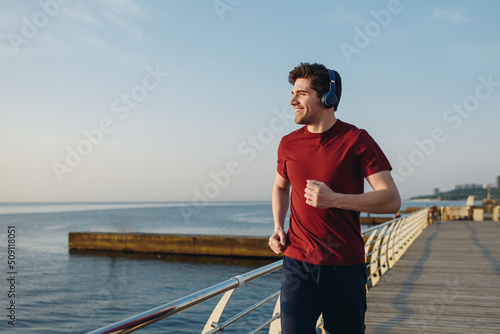 Young strong sporty athletic fit sportsman man wear sports clothes heapdhones listen music look aside warm up training at sunrise sun dawn over sea beach outdoor on pier seaside in summer day morning.