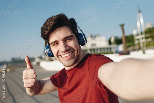Close up young strong sporty fit sportsman man in t-shirt headphones do selfie shot mobile cell phone show thumb up warm up train at sunrise sun dawn over sea beach outdoor on pier seaside in morning.