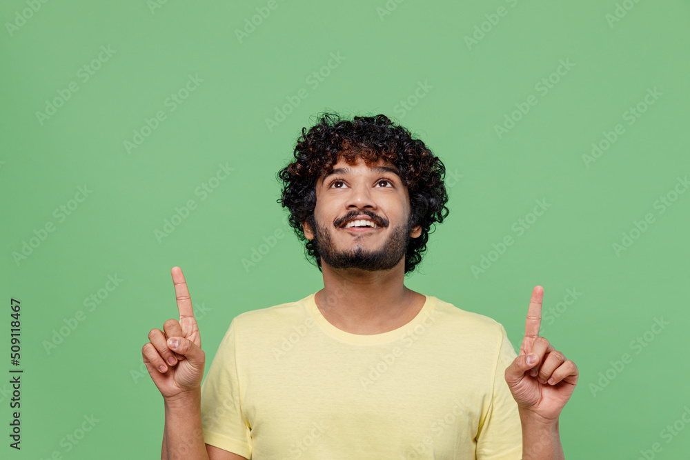 Young smiling happy Indian man 20s in yellow t-shirt point index finger overhead on workspace area mock up isolated on plain pastel light green background studio portrait. People lifestyle concept