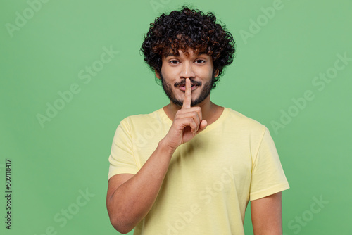 Young secret Indian man 20s wearing basic yellow t-shirt say hush be quiet with finger on lips shhh gesture isolated on plain pastel light green background studio portrait. People lifestyle concept. photo