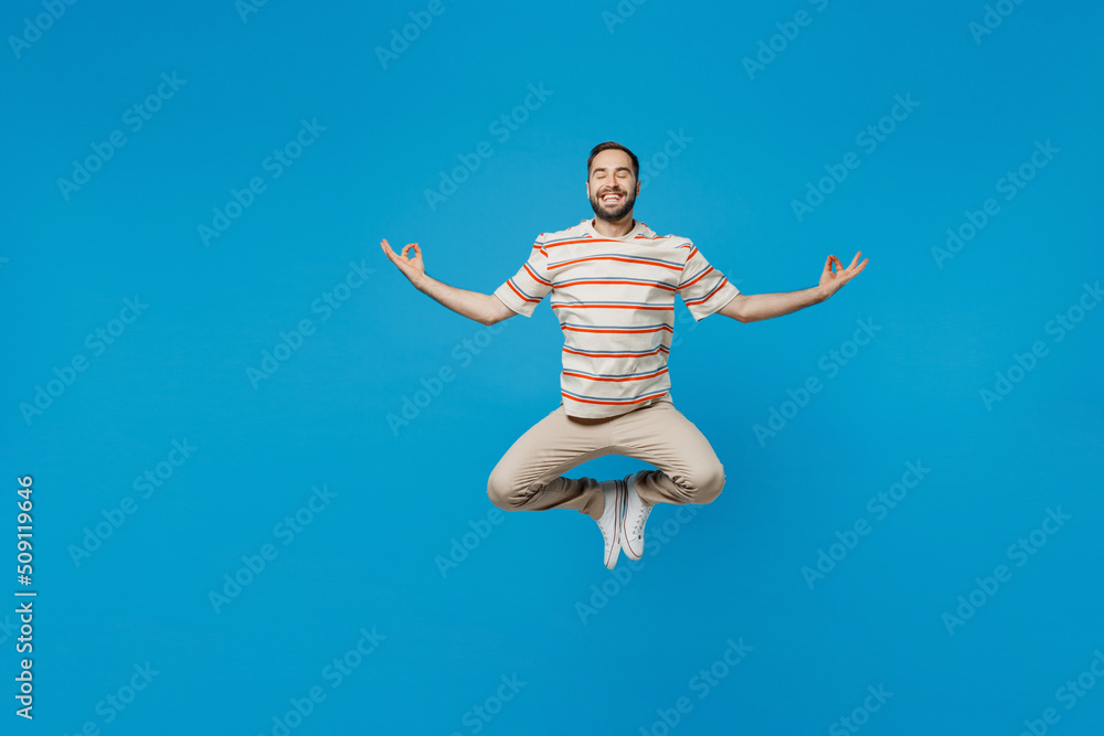 Full body young smiling man 20s wear orange striped t-shirt hold spreading hands in yoga om aum gesture relax meditate try to calm down levitating isolated on plain blue background studio portrait