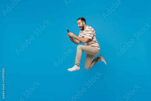 Wallpaper Mural Full body side view young excited fun cool overjoyed man 20s in orange striped t-shirt jump high hold use mobile cell phone isolated on plain blue background studio portrait. People lifestyle concept. Torontodigital.ca