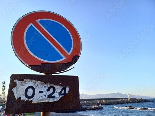 Segnale stradale di avvertimento, rotondo con bordo rosso e barrato su sfondo blu, che indica divieto di sosta dalle ore 0 alle ore 24, arruginito a causa della vicinanza al mare photo
