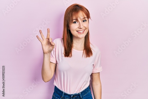 Redhead young woman wearing casual pink t shirt showing and pointing up with fingers number three while smiling confident and happy.