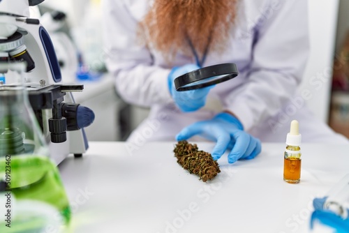 Fototapeta Naklejka Na Ścianę i Meble -  Young redhead man wearing scientist uniform holding marihuana weed and loupe at laboratory