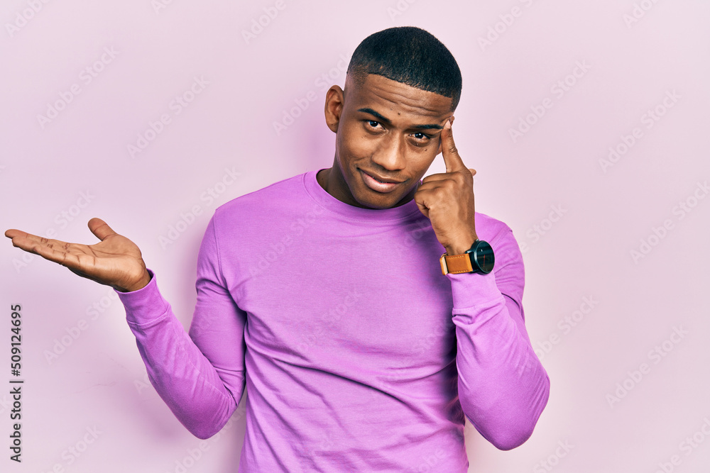 Young black man wearing casual pink sweater confused and annoyed with open palm showing copy space and pointing finger to forehead. think about it. Young black man wearing casual pink sweater confused and annoyed with open palm showing copy space and pointing finger to forehead. think about it.