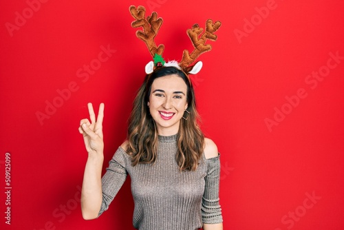 Young hispanic girl wearing deer christmas hat smiling with happy face winking at the camera doing victory sign. number two.