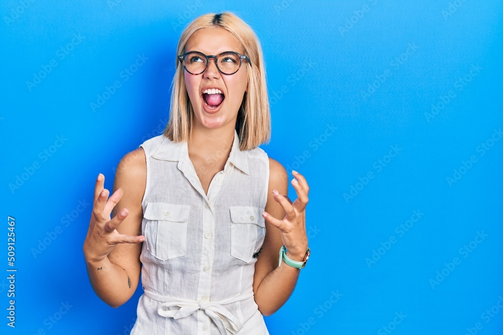 Beautiful blonde woman wearing glasses crazy and mad shouting and yelling with aggressive expression and arms raised. frustration concept. Beautiful blonde woman wearing glasses crazy and mad shouting and yelling with aggressive expression and arms raised. frustration concept.