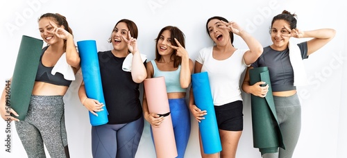 Group of women holding yoga mat standing over isolated background doing peace symbol with fingers over face, smiling cheerful showing victory